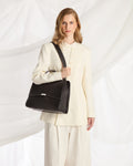 A woman wearing a cream-colored tailored suit carries a structured black leather shoulder bag with a silver-tone front clasp against a draped white fabric backdrop.