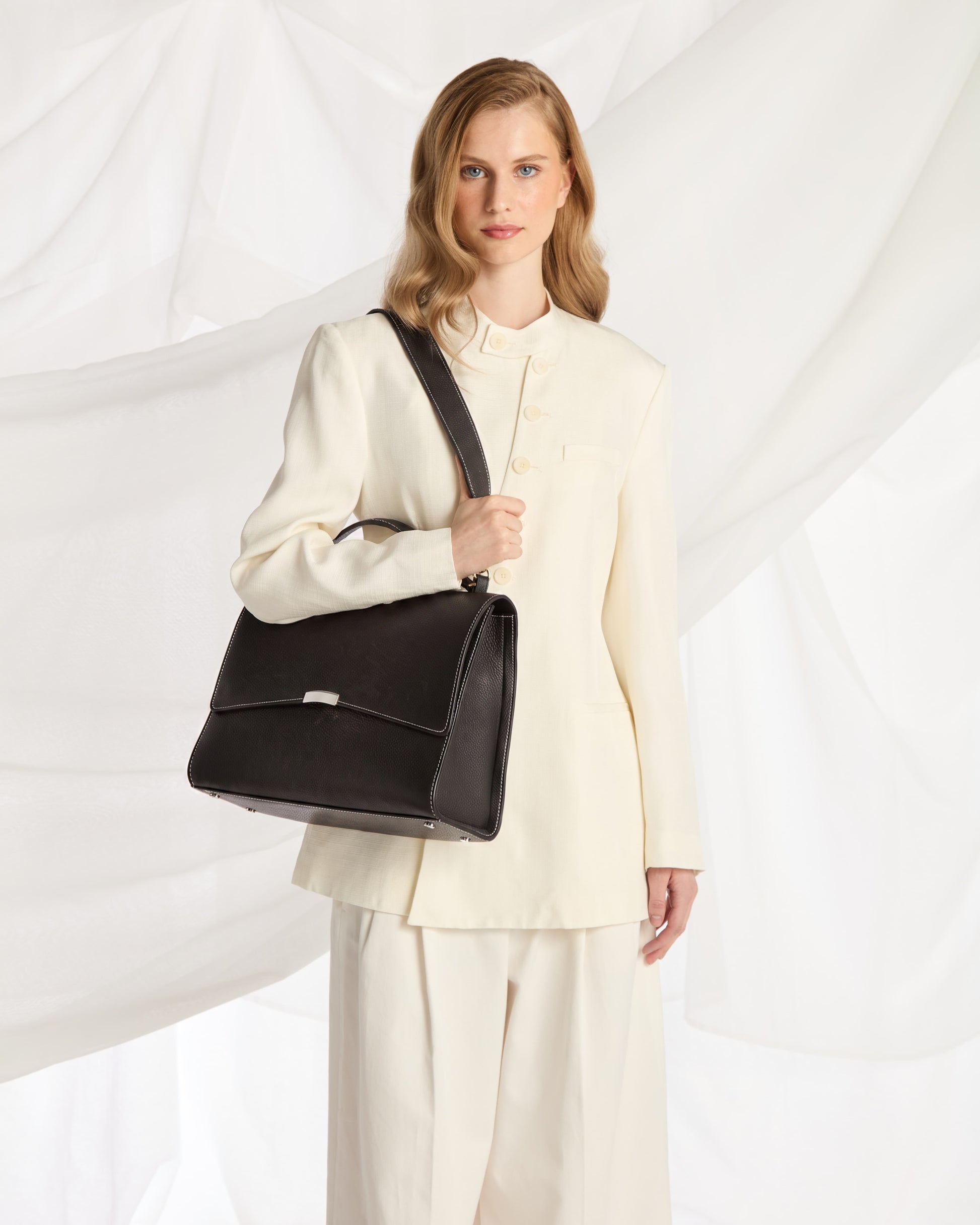 A woman wearing a cream-colored tailored suit carries a structured black leather shoulder bag with a silver-tone front clasp against a draped white fabric backdrop.