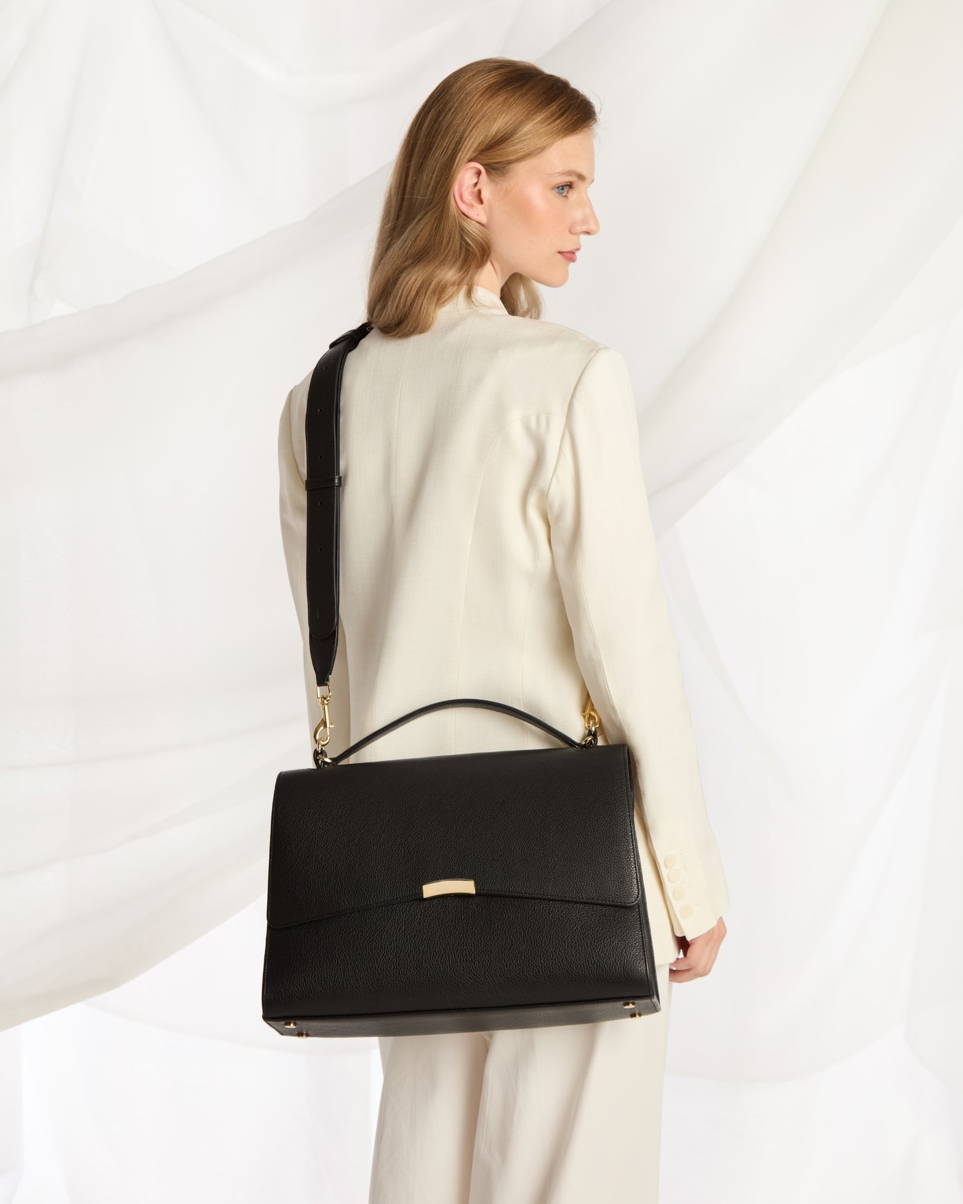 A woman in a cream-colored outfit is shown from the back, carrying a structured black leather cross body bag with gold-tone hardware against a white draped fabric background.