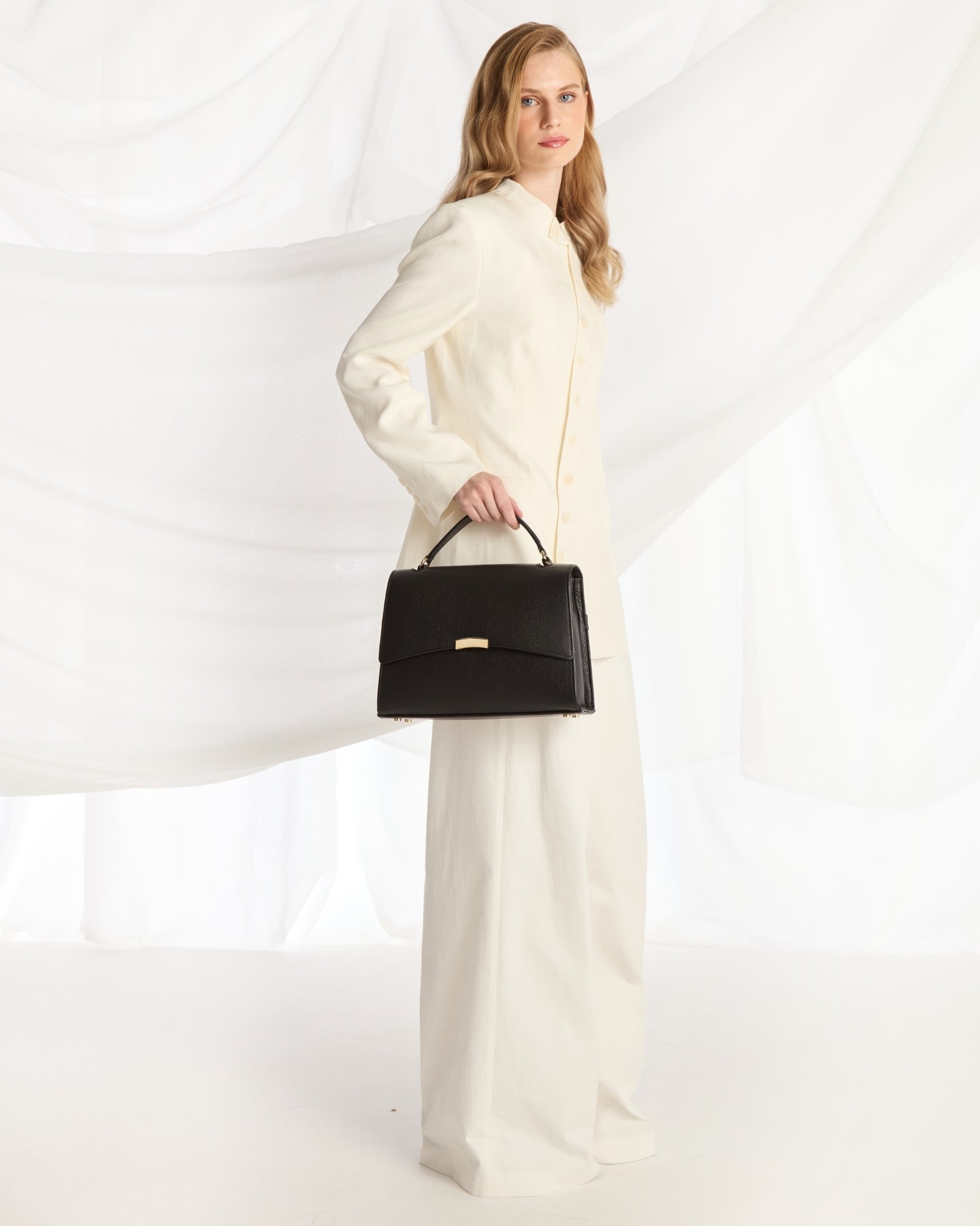 A woman modeling a long-sleeved cream-colored coat and matching wide-leg trousers, holding a structured black handbag with a gold-tone clasp against a white draped fabric backdrop.