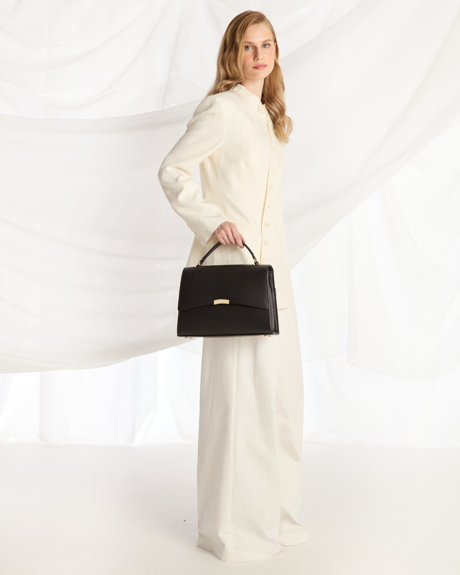 A woman modeling a long-sleeved cream-colored coat and matching wide-leg trousers, holding a structured black handbag with a gold-tone clasp against a white draped fabric backdrop.
