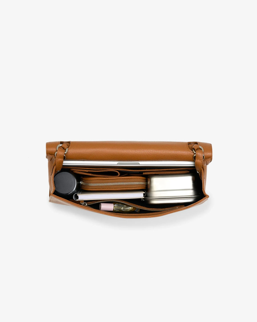 A tan leather travel pouch shown open from a top-down view, containing travel accessories such as a silver metal can, a black travel mug, and a small pink item, on a light gray background.
