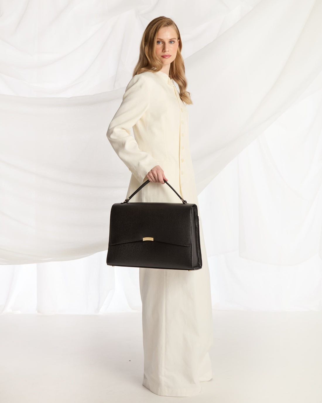 A woman wearing a cream-colored tailored suit and holding a black leather shoulder bag with a strap, posed against a white draped fabric background.