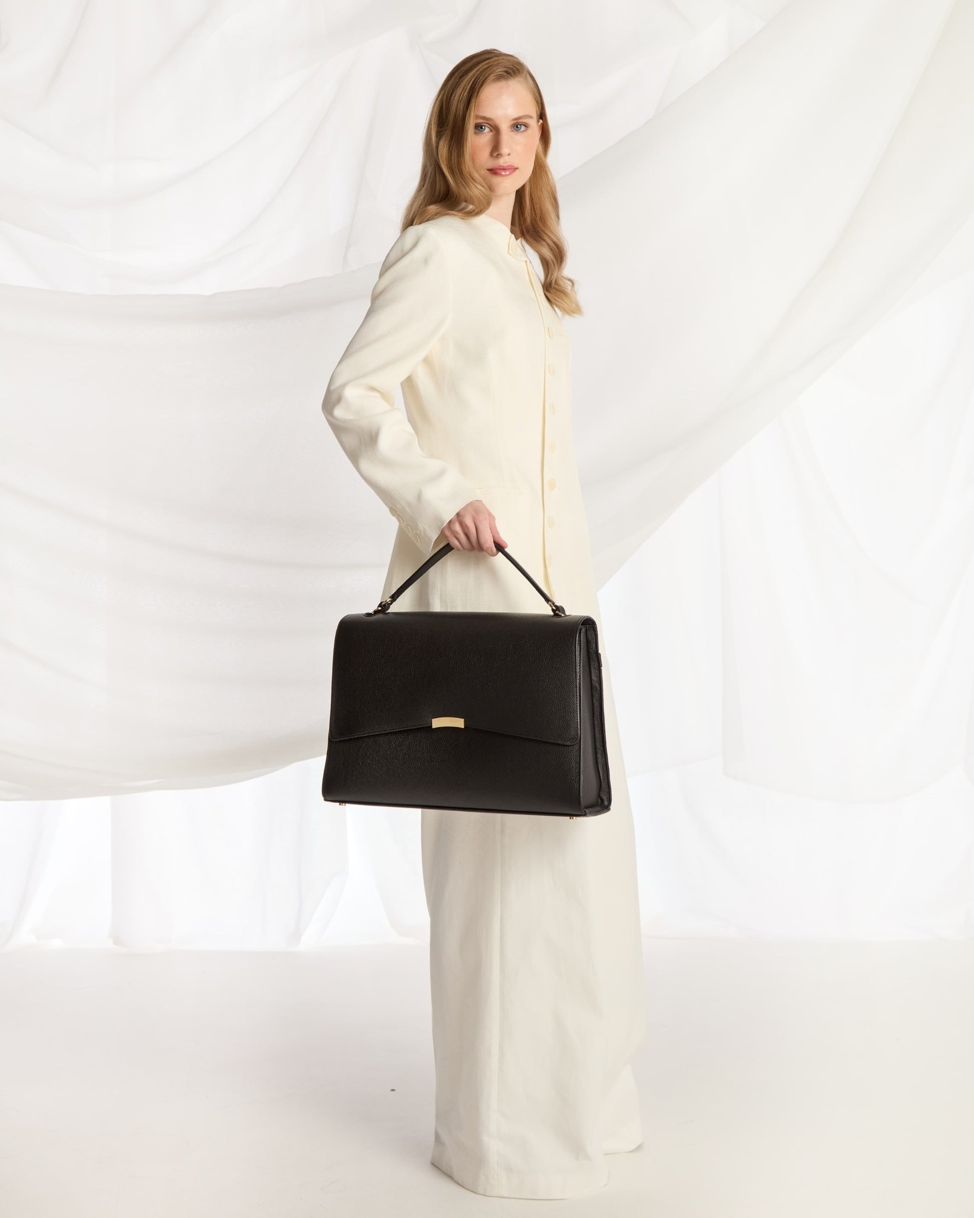 A woman wearing a cream-colored tailored suit and holding a black leather shoulder bag with a strap, posed against a white draped fabric background.