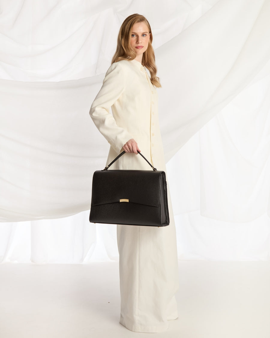 A woman wearing a cream-colored tailored suit and holding a black leather shoulder bag with a strap, posed against a white draped fabric background.