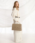 A woman in a flowing cream-colored outfit holds a structured taupe leather handbag with a top handle, posed against a draped white fabric backdrop.
