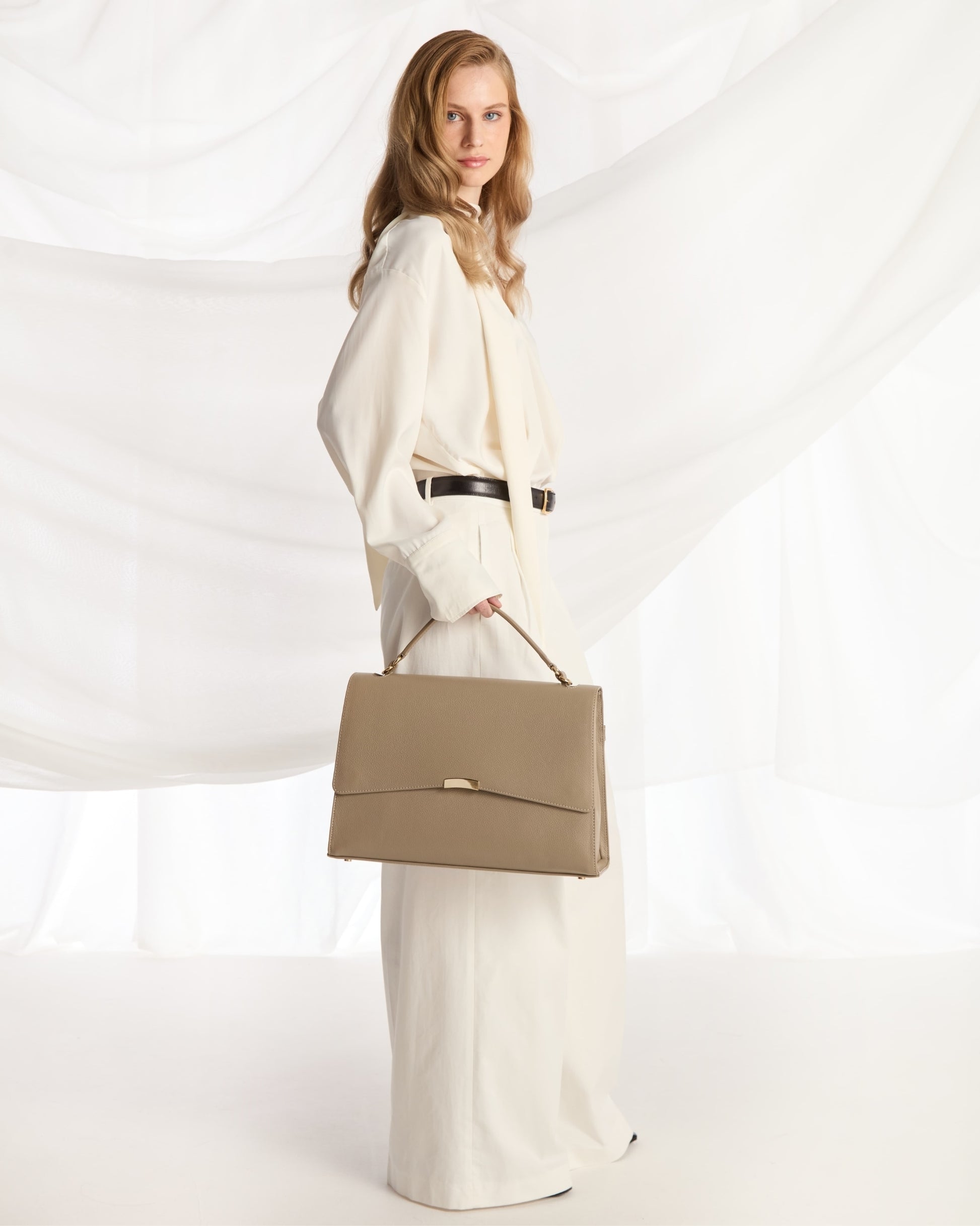A woman in a flowing cream-colored outfit holds a structured taupe leather handbag with a top handle, posed against a draped white fabric backdrop.