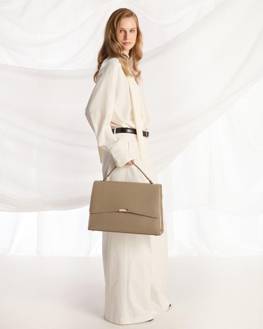A woman in a flowing cream-colored outfit holds a structured taupe leather handbag with a top handle, posed against a draped white fabric backdrop.