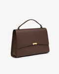 A dark brown textured leather handbag with a top handle and a small gold-tone metal accent on the front flap, shown against a plain light gray background.