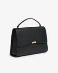 A black leather top-handle handbag with a gold-tone metal accent on the front flap, shown from a three-quarters angle against a plain white background.
