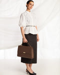 A woman stands in a studio wearing a white puff-sleeve blouse with a thin black belt, a black midi skirt, and black heels, while holding a structured brown handbag with a top handle and small gold-tone hardware.