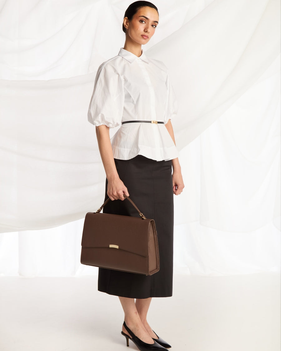 A woman stands in a studio wearing a white puff-sleeve blouse with a thin black belt, a black midi skirt, and black heels, while holding a structured brown handbag with a top handle and small gold-tone hardware.