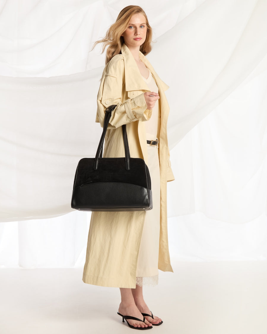 A woman modeling a long cream trench coat and matching midi dress, carrying a large black tote bag, against a white draped fabric backdrop.