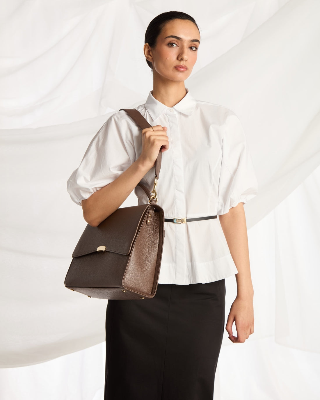 A woman wearing a white blouse and black skirt carries a brown leather shoulder bag with a gold-tone clasp and strap against a draped white fabric background.
