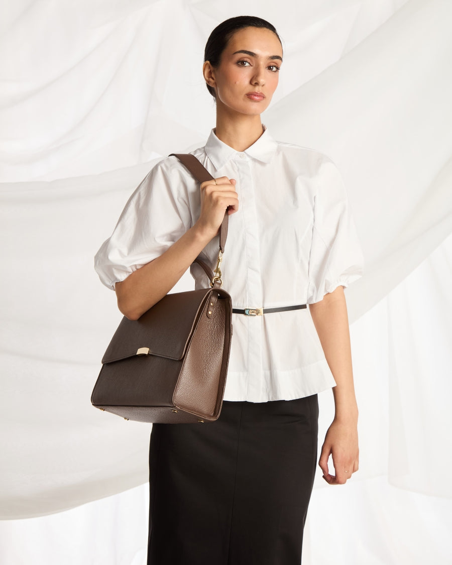 A woman wearing a white blouse and black skirt carries a brown leather shoulder bag with a gold-tone clasp and strap against a draped white fabric background.