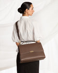 A woman wearing a white blouse and black skirt is shown from the back while carrying a brown leather shoulder bag with a top handle and flap closure against a white draped fabric background.