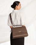 A woman wearing a white blouse and black skirt is shown from the back while carrying a brown leather cross body bag with a top handle and flap closure against a white draped fabric background.