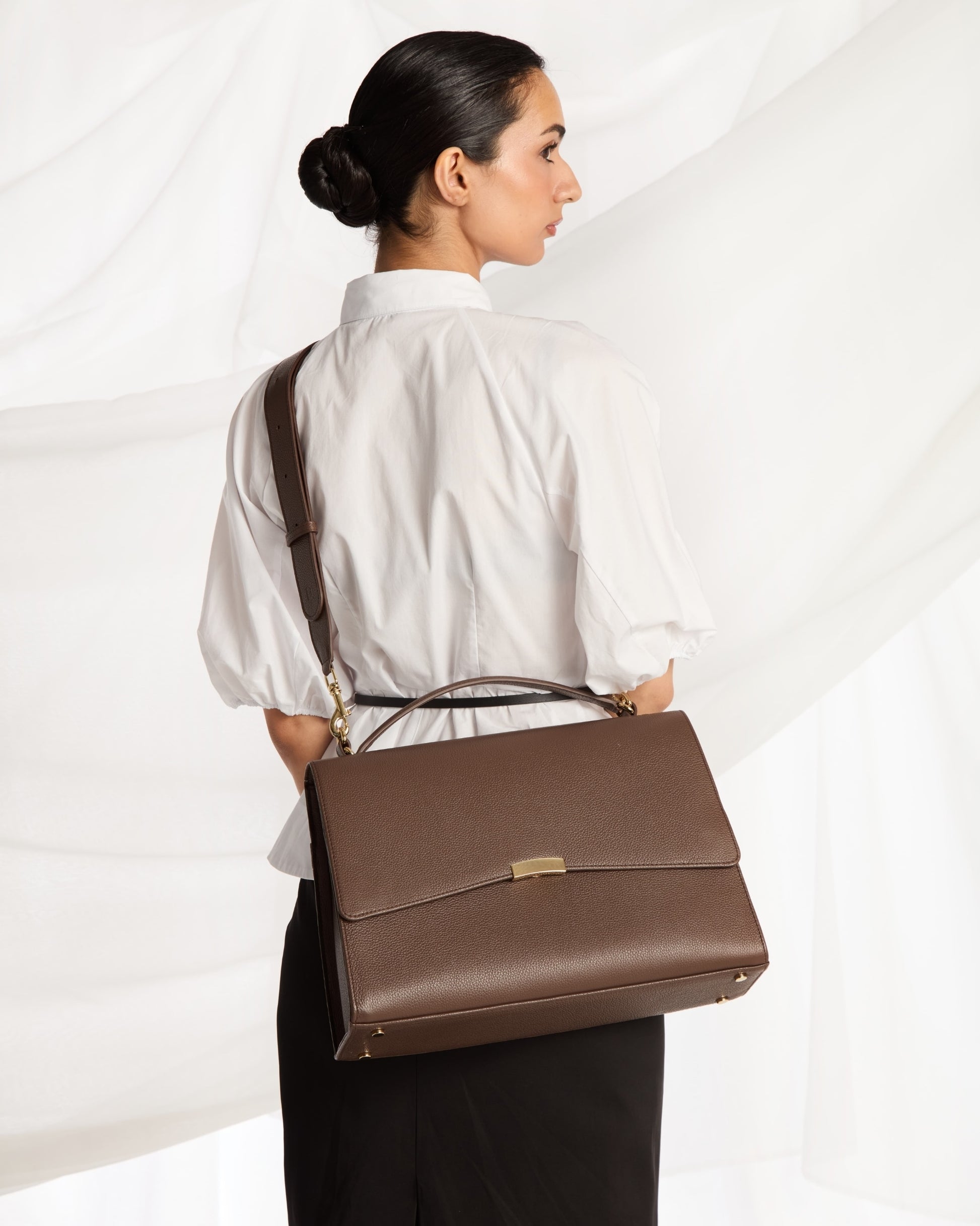 A woman wearing a white blouse and black skirt is shown from the back while carrying a brown leather cross body bag with a top handle and flap closure against a white draped fabric background.
