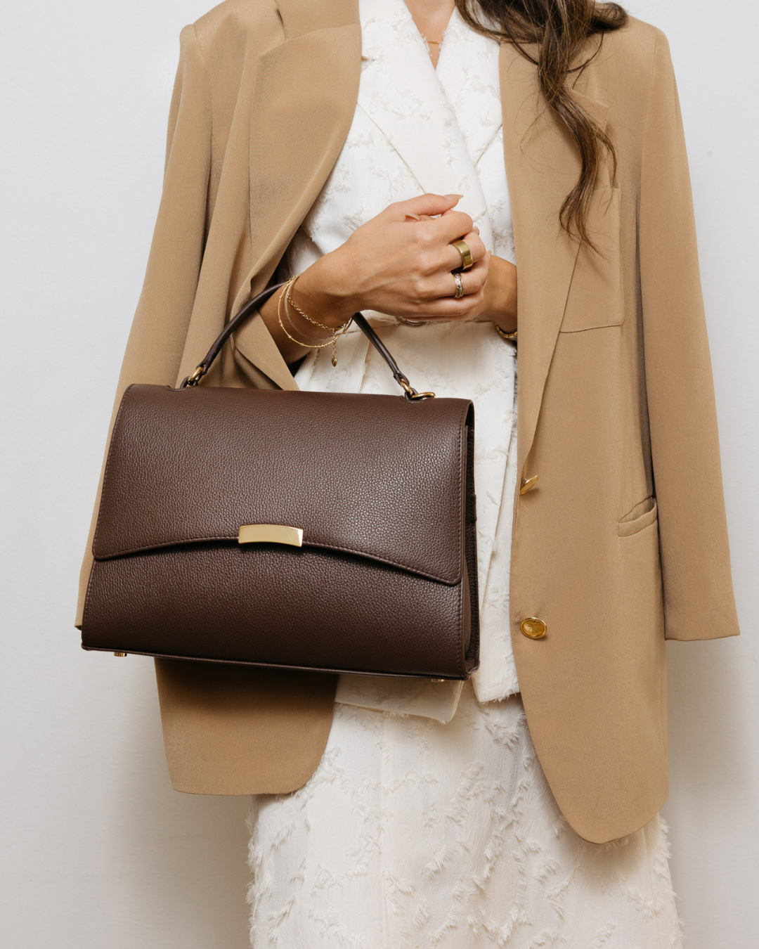 A structured dark brown leather handbag with a top handle and a small gold-tone metal accent on the front flap, shown against a plain light gray background.