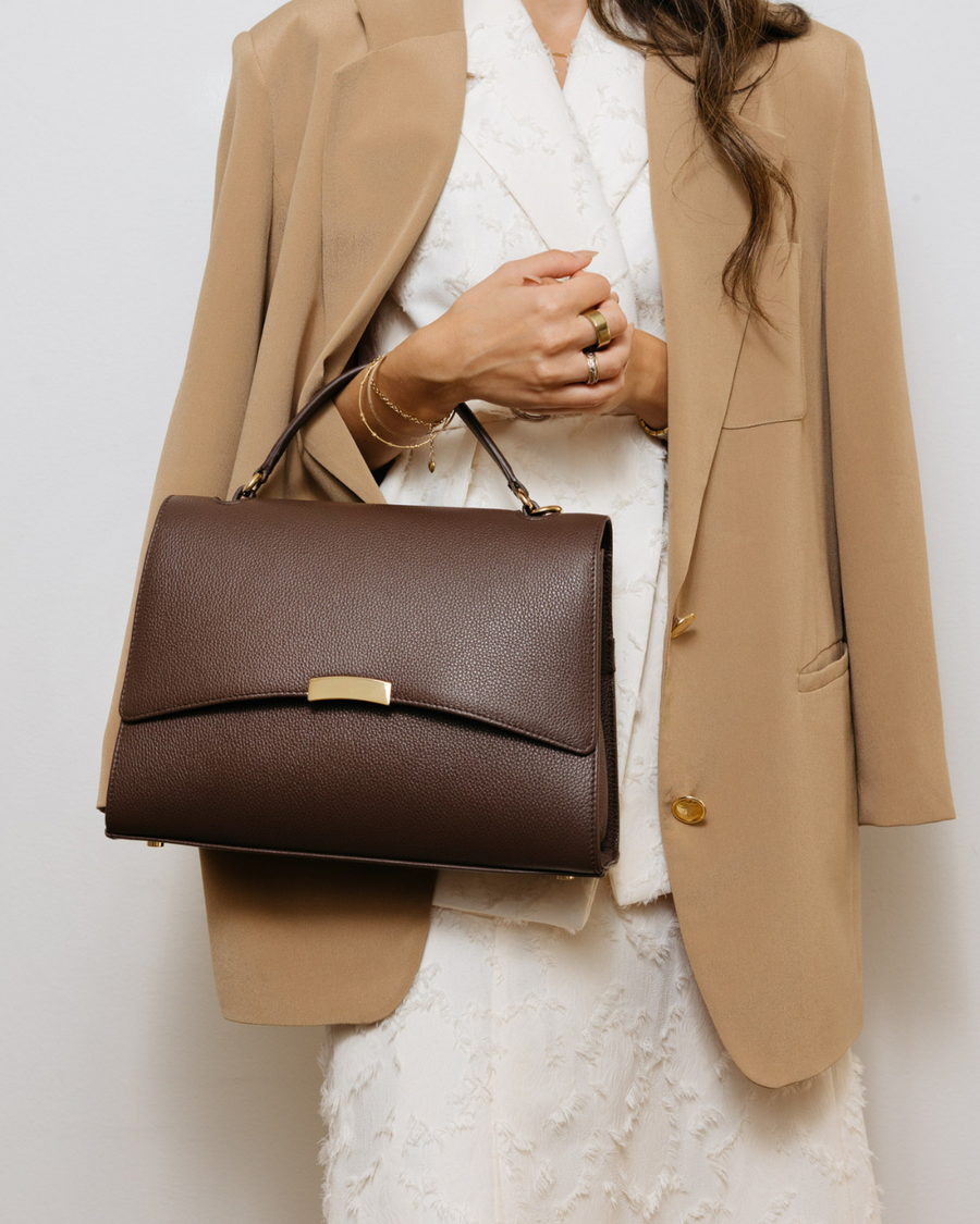 Person wearing a beige blazer and white lace top, holding a brown handbag against a light background