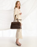 A woman modeling a large dark brown leather tote bag, wearing a cream-colored dress and black sandals, set against a white draped fabric background.