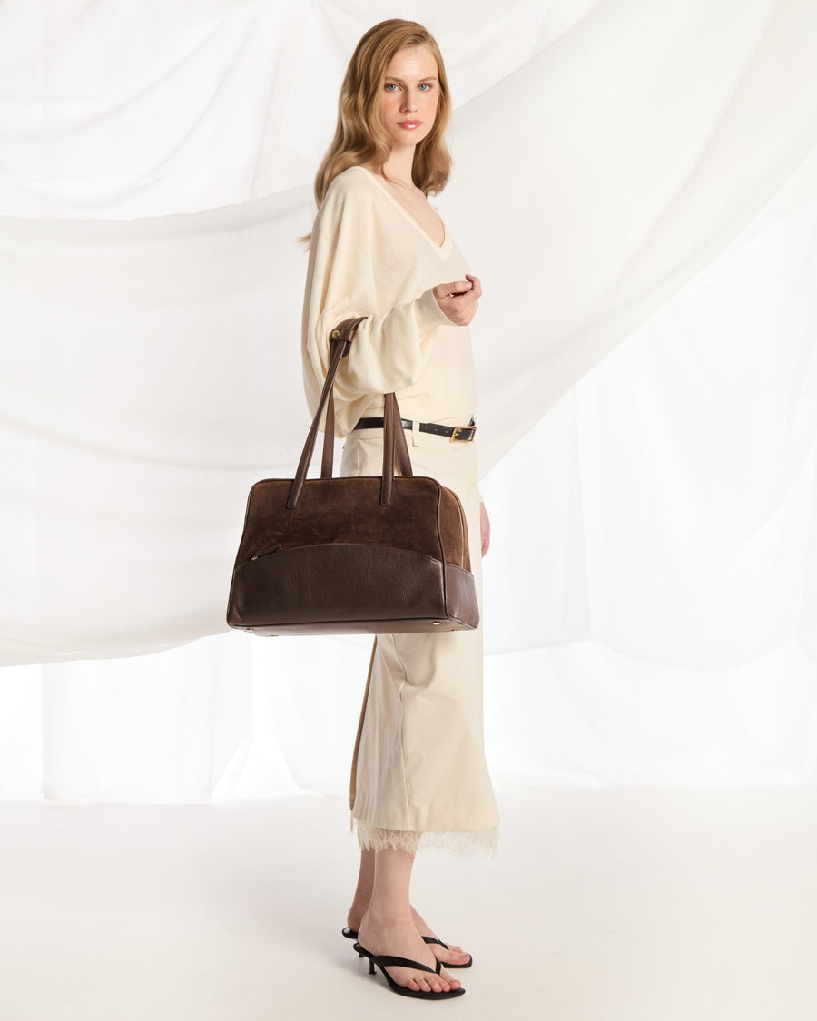 A woman modeling a large dark brown leather tote bag, wearing a cream-colored dress and black sandals, set against a white draped fabric background.