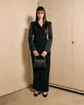 A woman stands in a minimalist indoor setting wearing a black tailored blazer and matching wide-leg trousers, holding a structured black handbag, with black pointed flats and orange-tinted sunglasses.