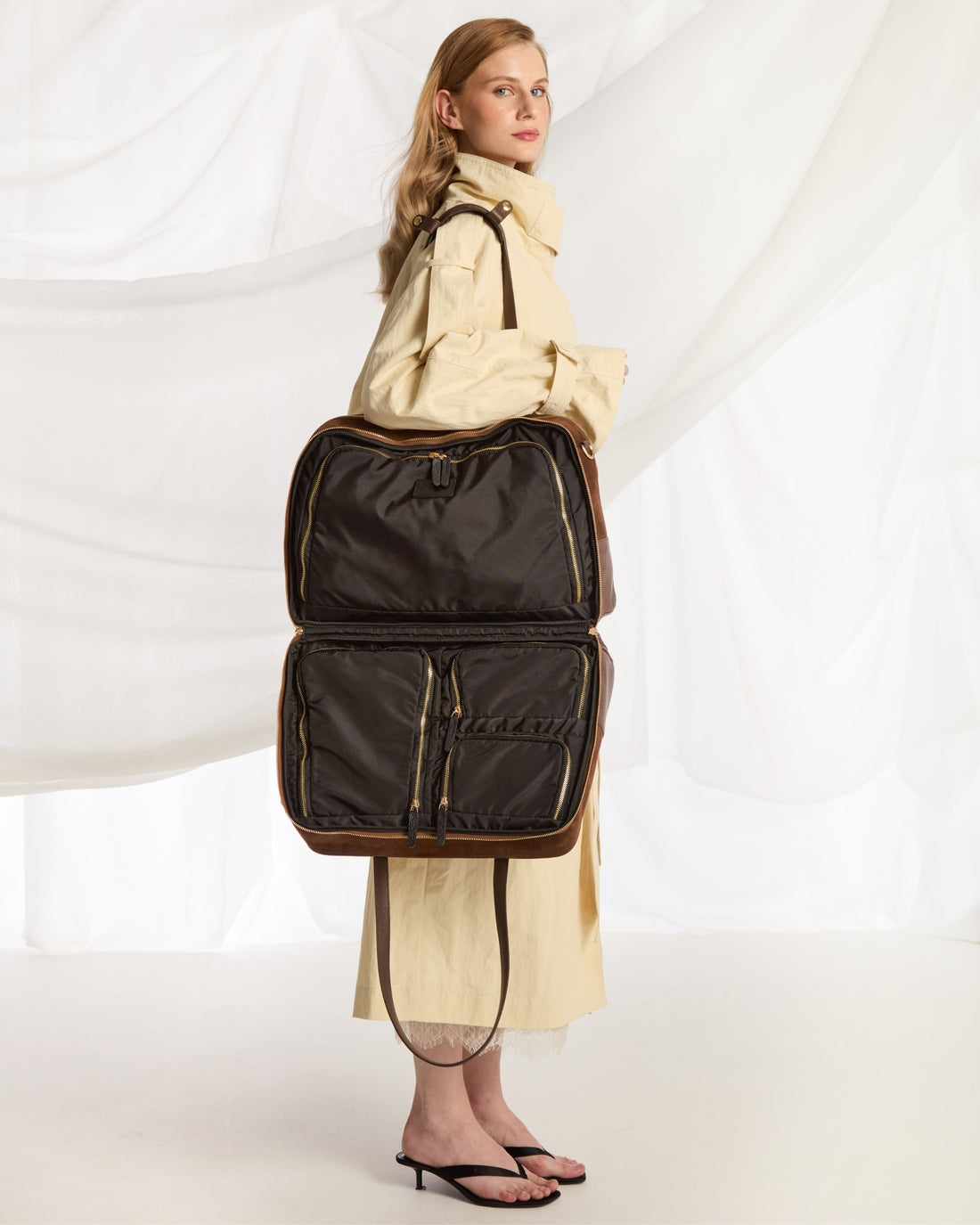 A model in a cream trench coat and sandals carries a large black nylon suitcase-style bag with brown trim and gold-toned zipper details against a white draped studio backdrop.