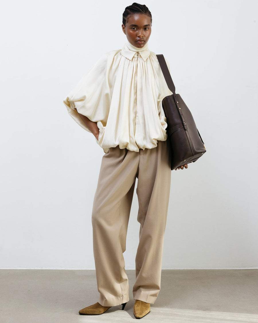 Woman wearing a white blouse and beige pants holding a brown bag against a plain background