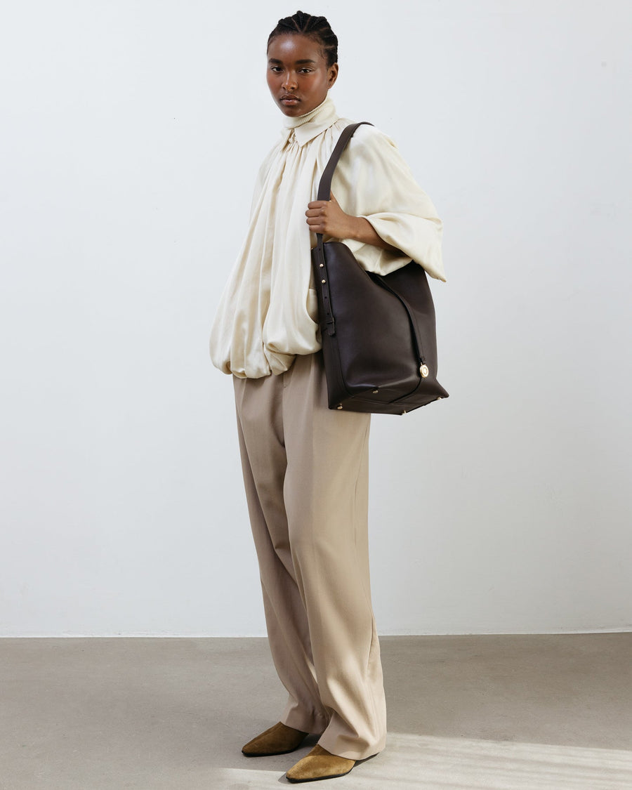 Person wearing a white shirt and beige pants holding a brown leather bag against a plain background