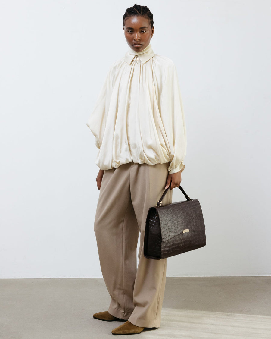 Person wearing a cream blouse and beige pants holding a brown handbag against a white background