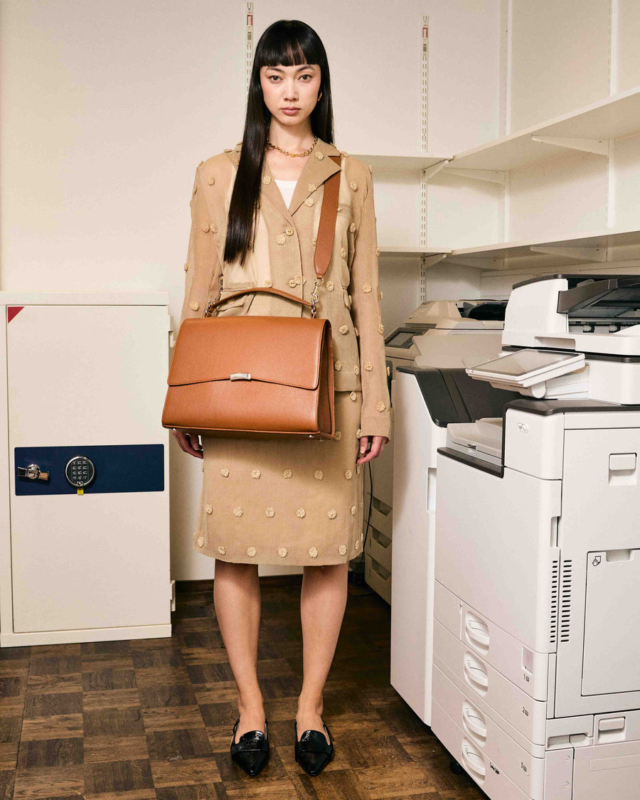 A woman wearing a tan briefcase with metal hardware, carrying it with a cross body strap.