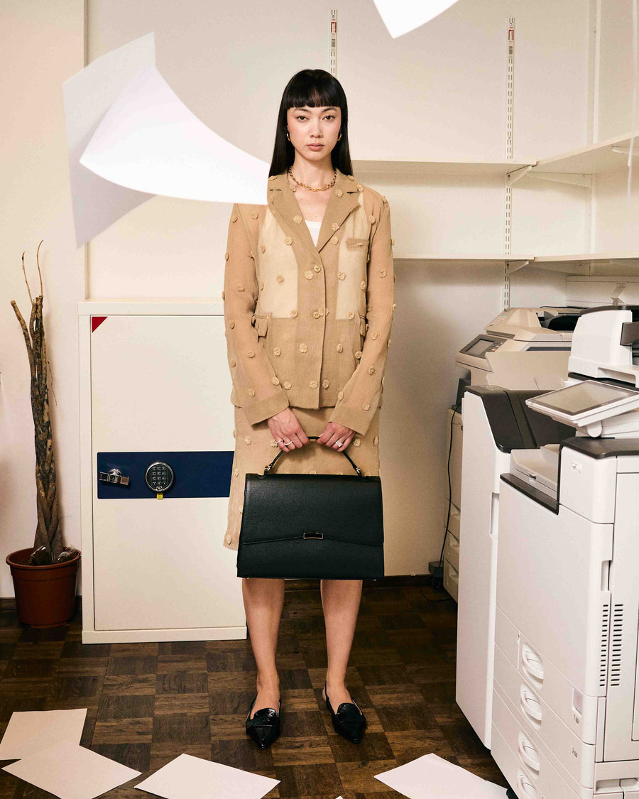 A woman in an office setting, wearing a beige outfit and holding a black leather briefcase with silver hardware.