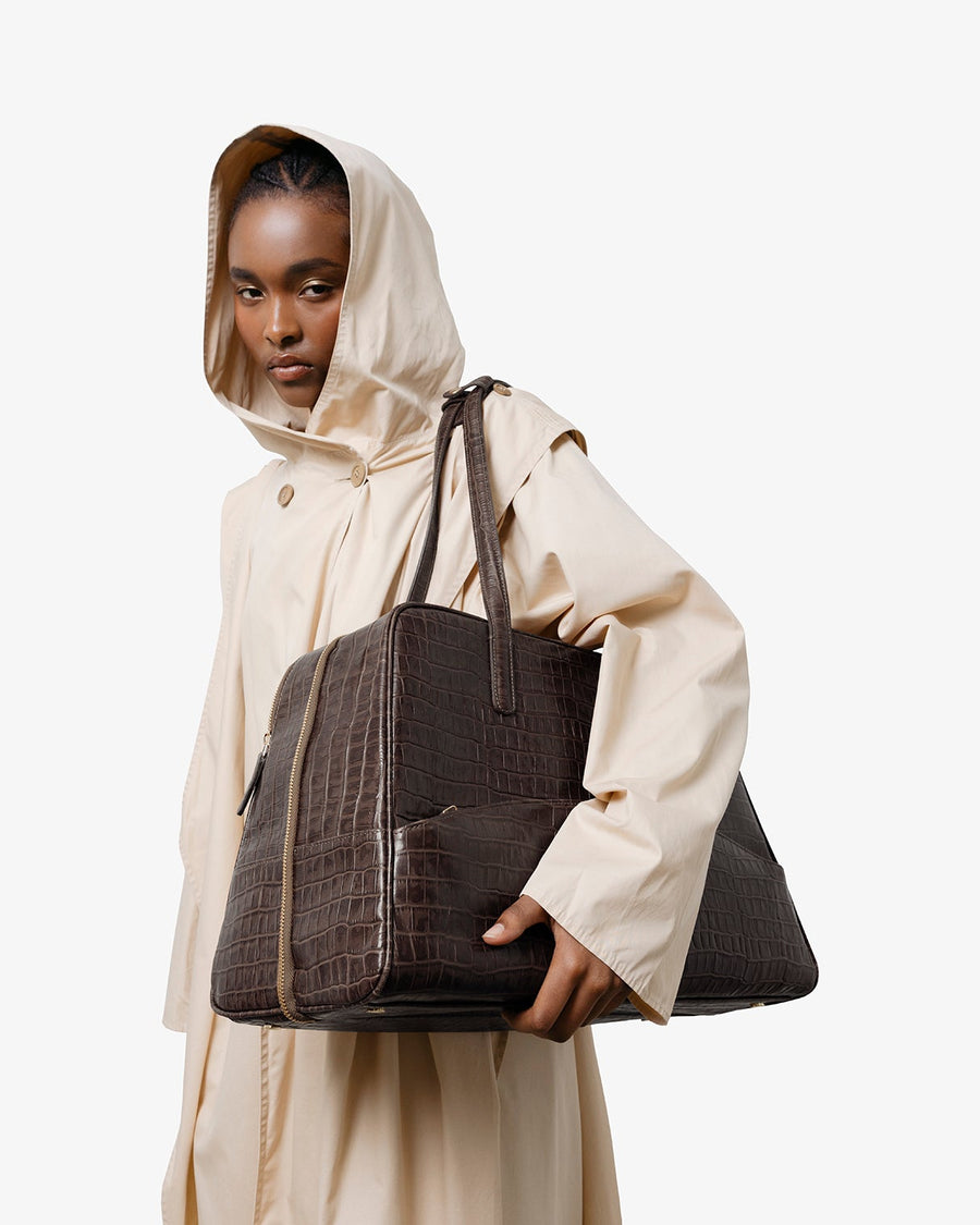 Person wearing a beige raincoat holding a textured brown leather bag on a white background