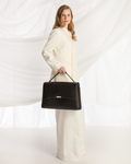A woman wearing a cream-colored long coat and wide-leg trousers holds a structured black leather briefcase-style handbag with a top handle, posed against a draped white fabric backdrop.