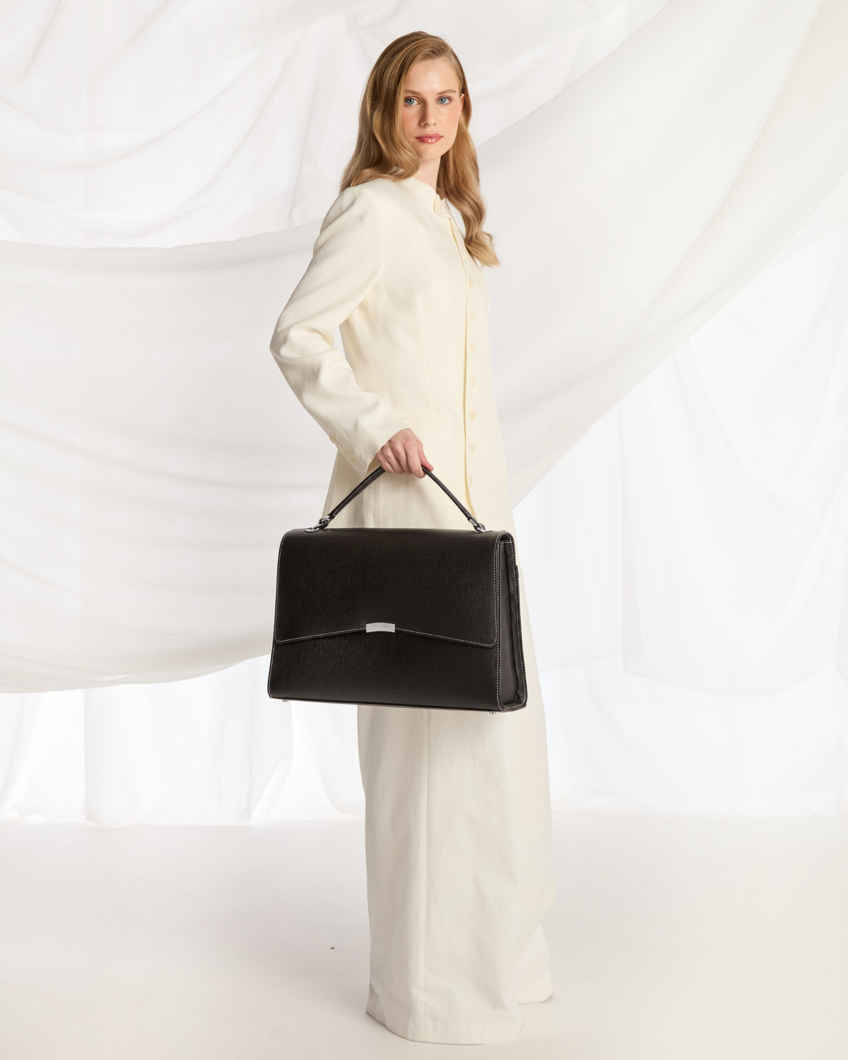 A woman wearing a cream-colored long coat and wide-leg trousers holds a structured black leather briefcase-style handbag with a top handle, posed against a draped white fabric backdrop.