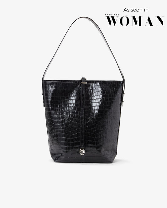 A black crocodile-embossed leather shoulder bag with a single strap, shown against a light gray background with 'As seen in Emirates WOMAN' text in the upper right.