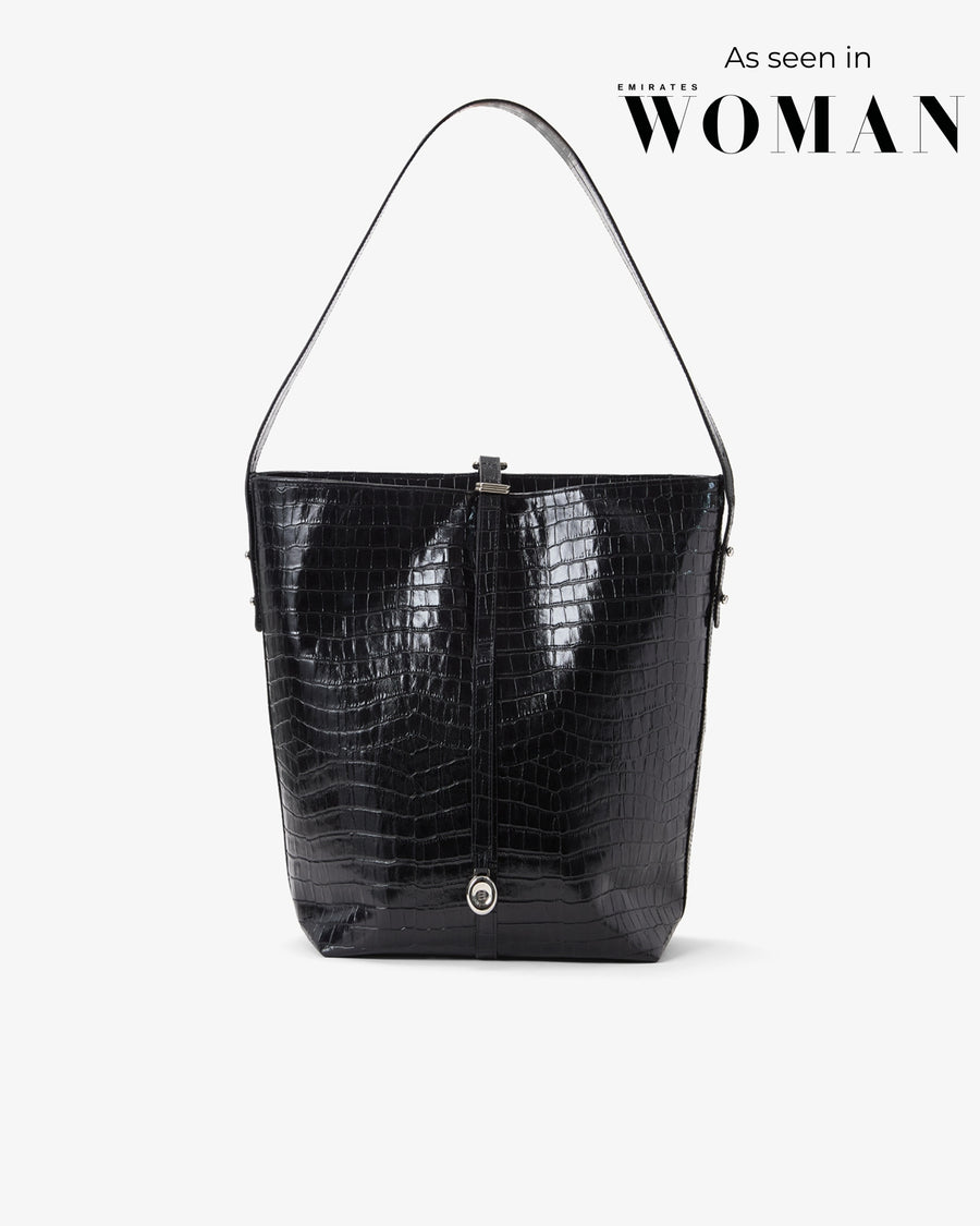 A black crocodile-embossed leather shoulder bag with a single strap, shown against a light gray background with 'As seen in Emirates WOMAN' text in the upper right.