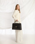 A woman in a long cream-colored dress holds a structured black leather briefcase-style bag with a top handle and a small gold front accent, posed against a draped white fabric backdrop.