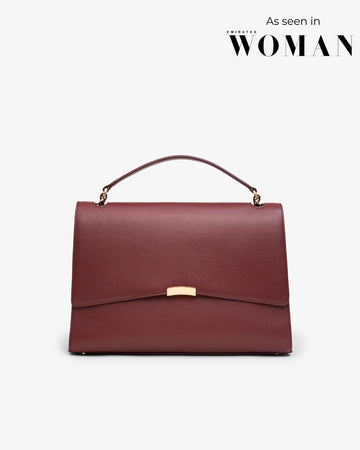A structured top-handle handbag in burgundy textured leather with a gold-tone clasp and hardware, shown against a light gray background with 'As seen in WOMAN' text in the top right corner.
