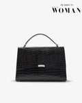 A black crocodile-embossed leather handbag with a top handle and a small silver metal accent, shown against a light gray background with 'As seen in WOMAN' text in the upper right.