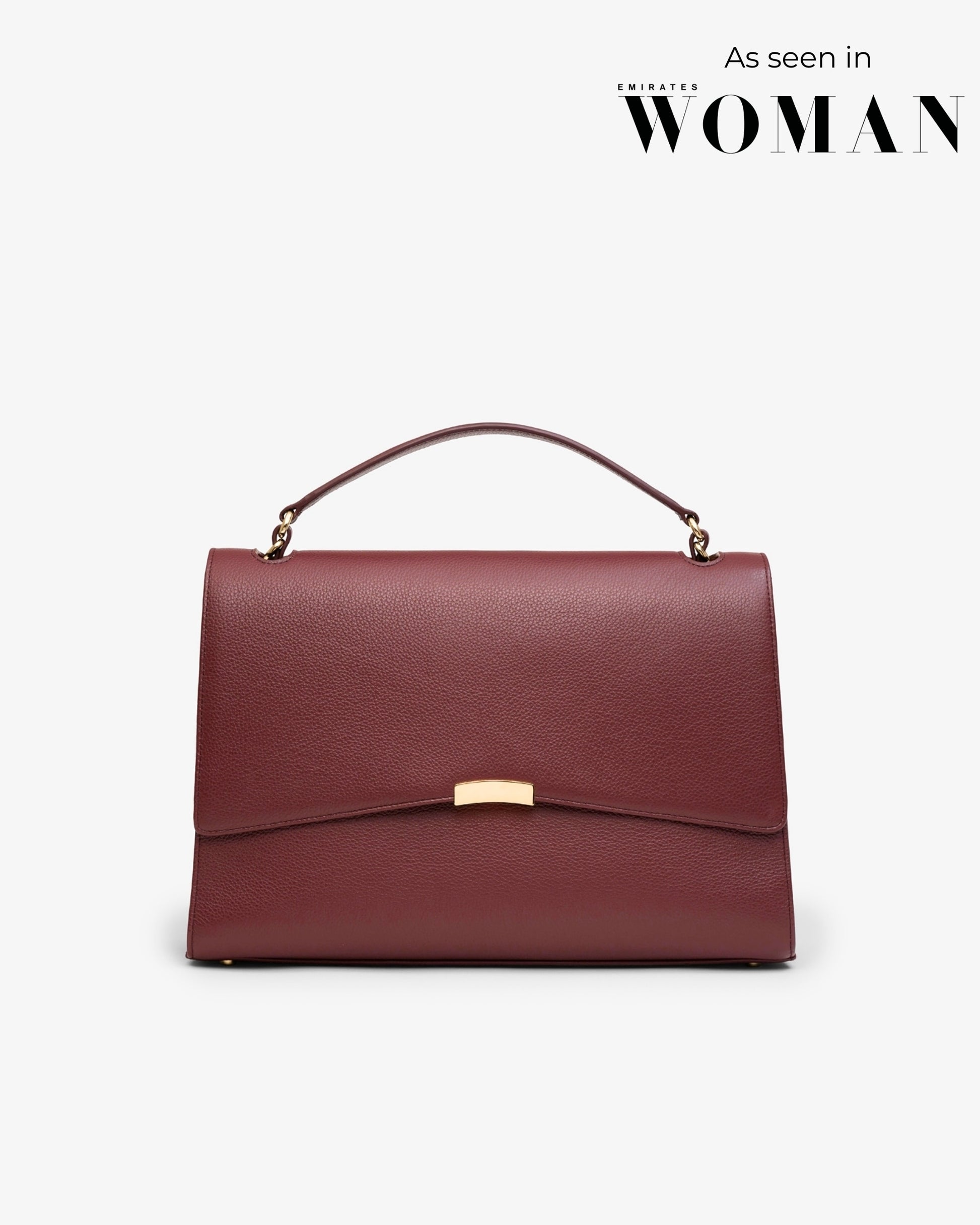 A structured top-handle handbag in burgundy textured leather with a gold-tone clasp and hardware, shown against a light gray background with 'As seen in EMIRATES WOMAN' text in the top right corner.