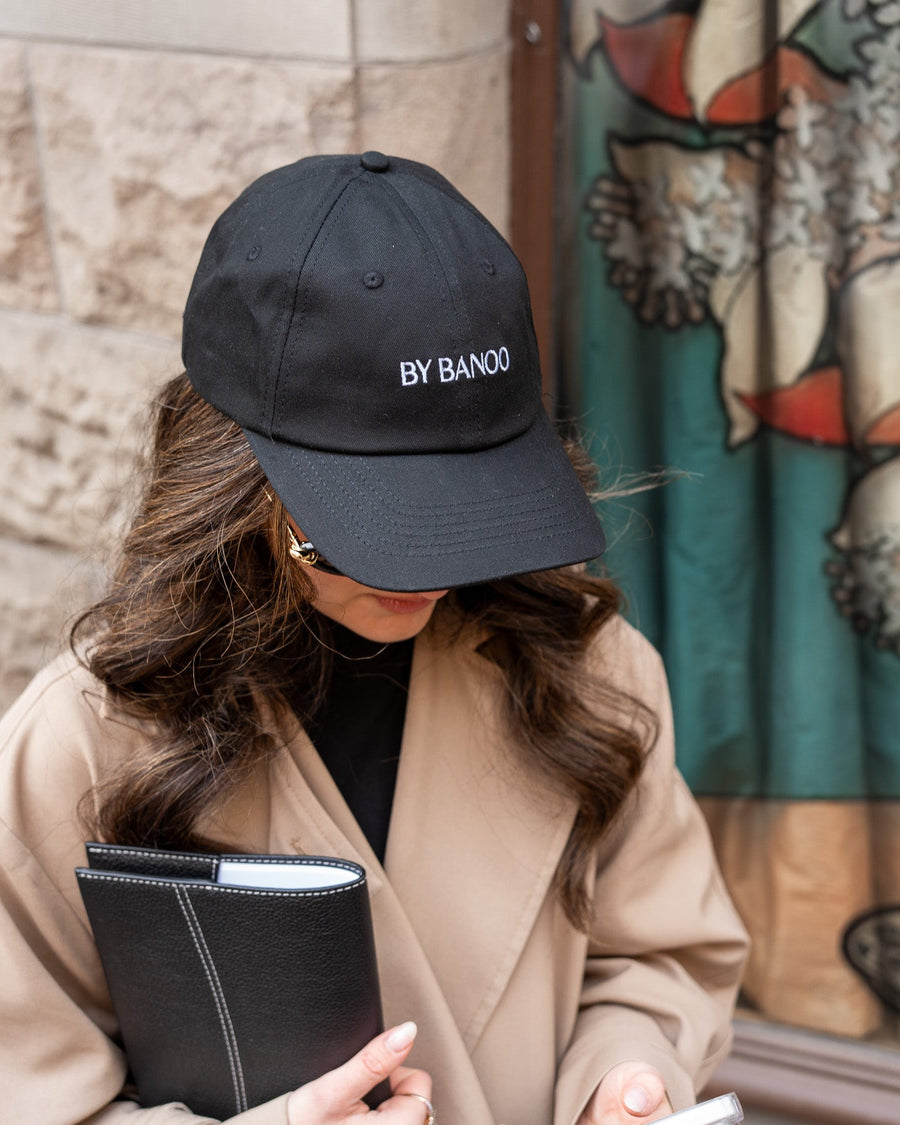 Person wearing a black cap with 'BY BANCO' logo, holding a phone and notebook, standing in front of a colorful mural.