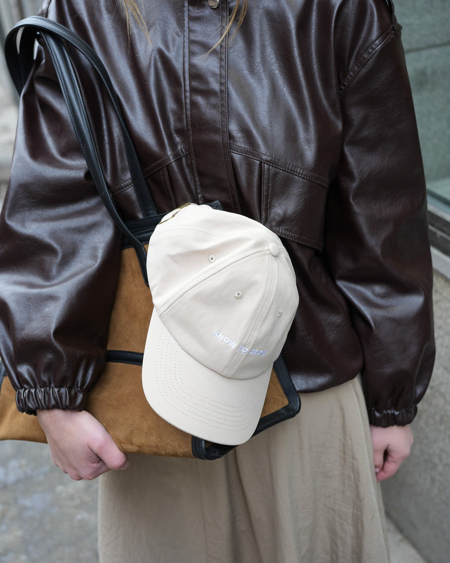 Person wearing a brown leather jacket holding a beige cap and brown bag.