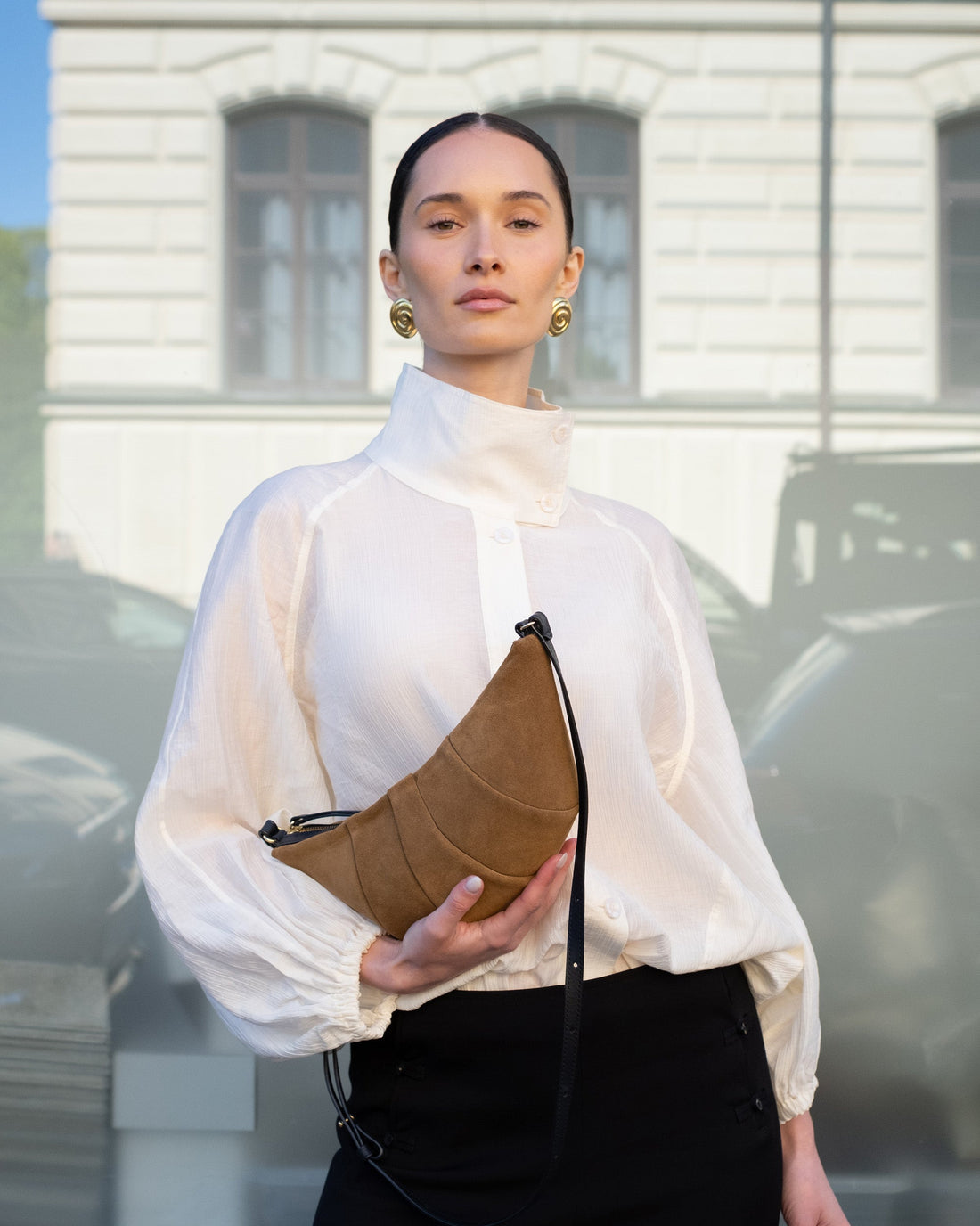 A brown suede crescent-shaped shoulder bag with a black leather strap, displayed against a clean white background.