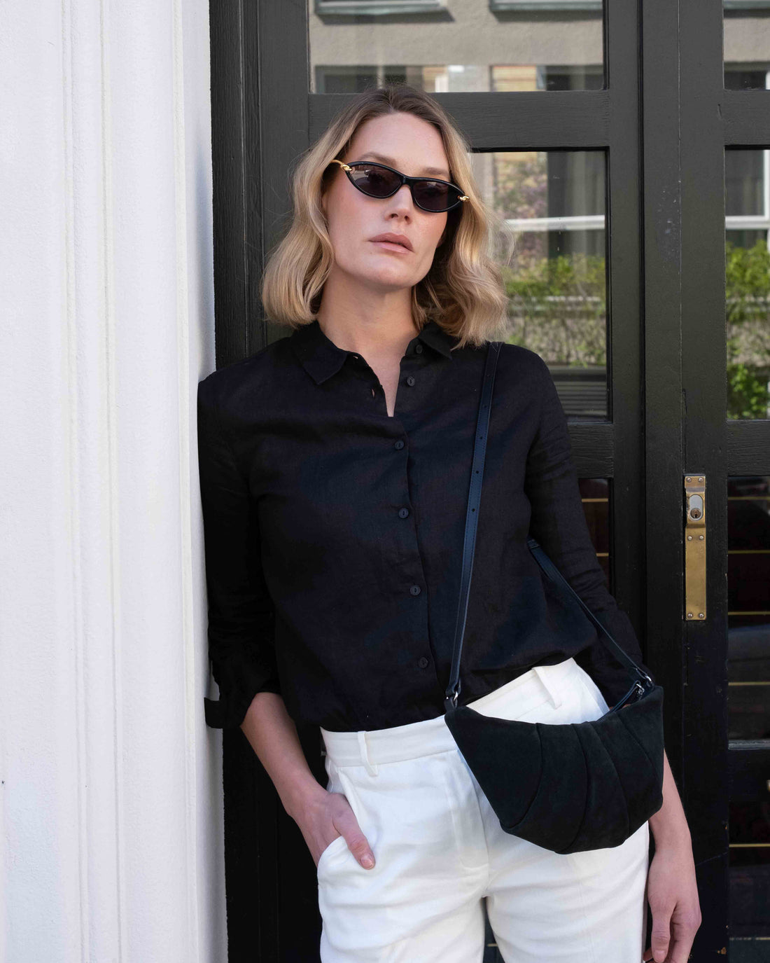 Woman wearing sunglasses, a black jacket and a suede shoulder bag, leaning against a building.