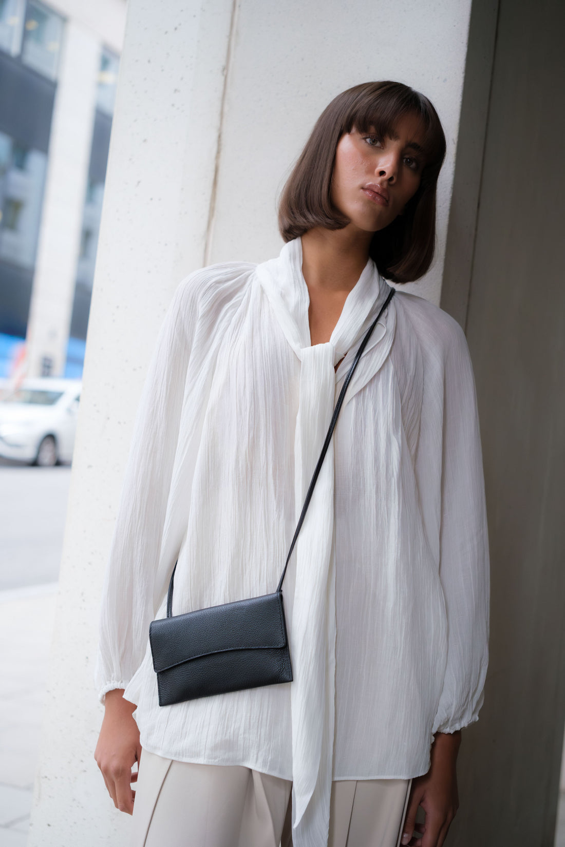 A minimalist black leather crossbody bag with a slim, long strap, shown against a plain light gray background.