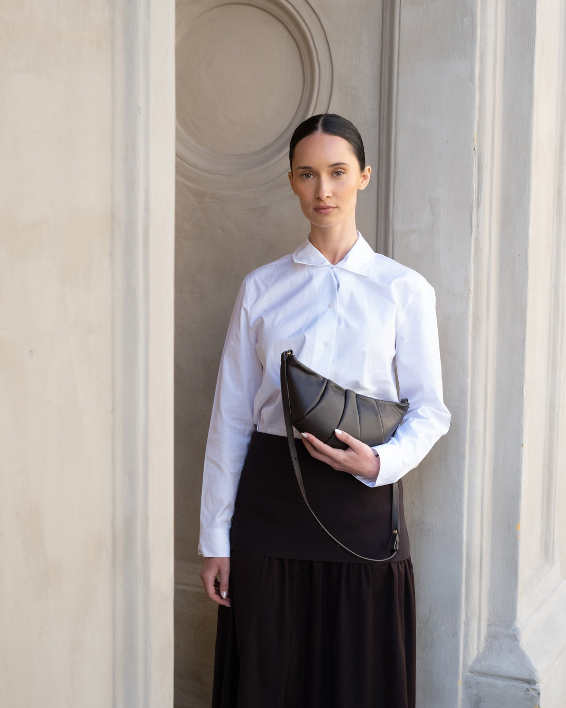 A dark brown leather crescent shoulder bag with a pleated front design and a thin adjustable shoulder strap, shown against a plain white background.