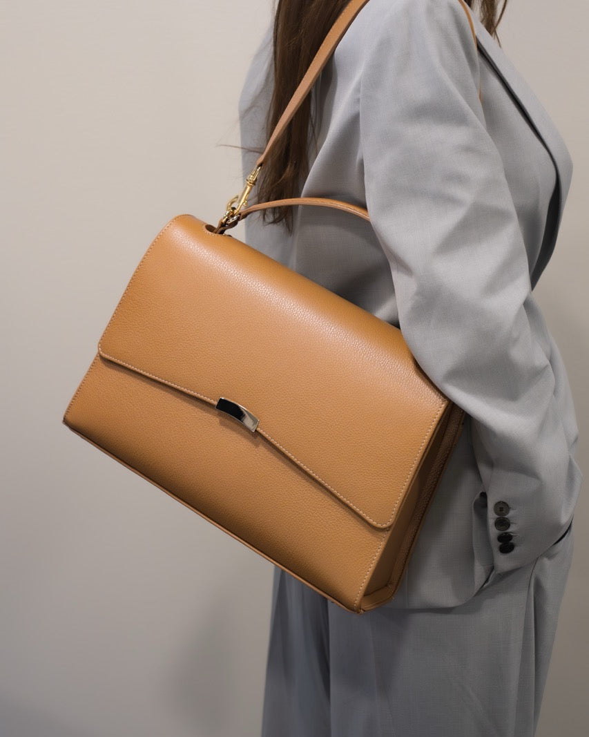 Person wearing a light gray coat holding a tan leather handbag against a plain background