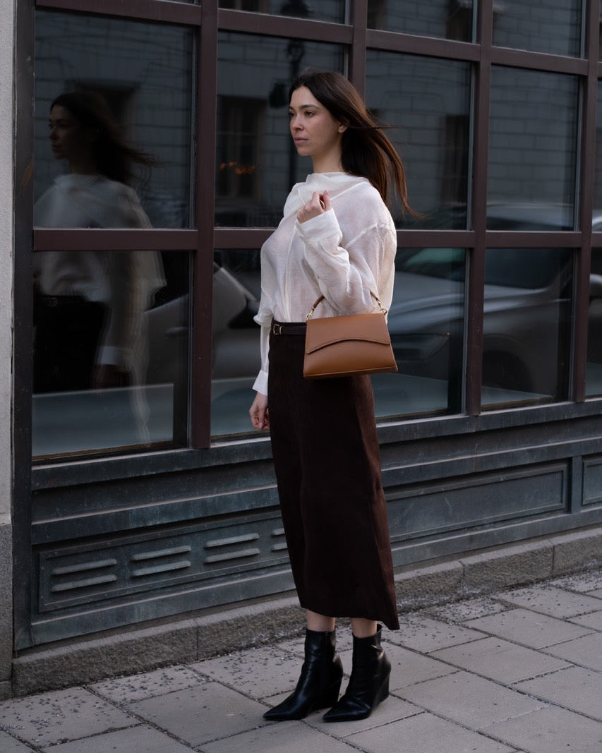 Woman standing on a street corner wearing a white blouse and brown skirt, holding a brown handbag.